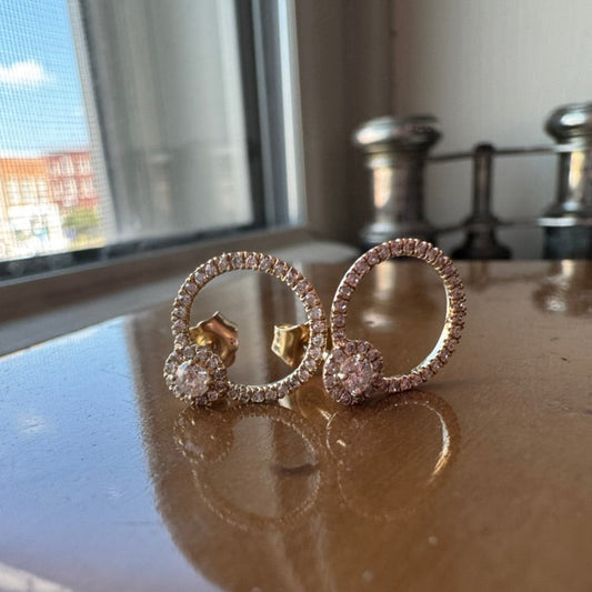 Yellow gold earrings with diamonds on a reflective surface near a window.