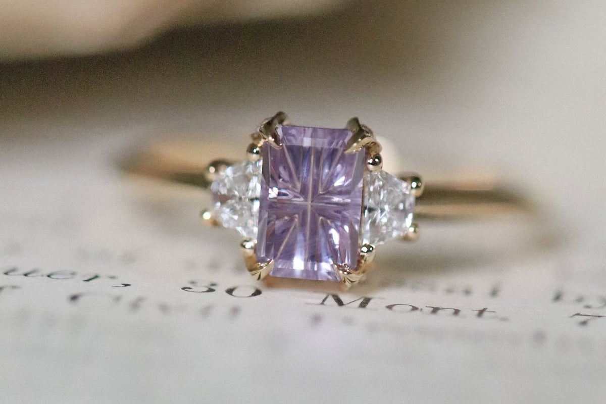 A yellow gold ring featuring a rectangular lavender amethyst center stone with two clear round side stones, resting on an open book.
