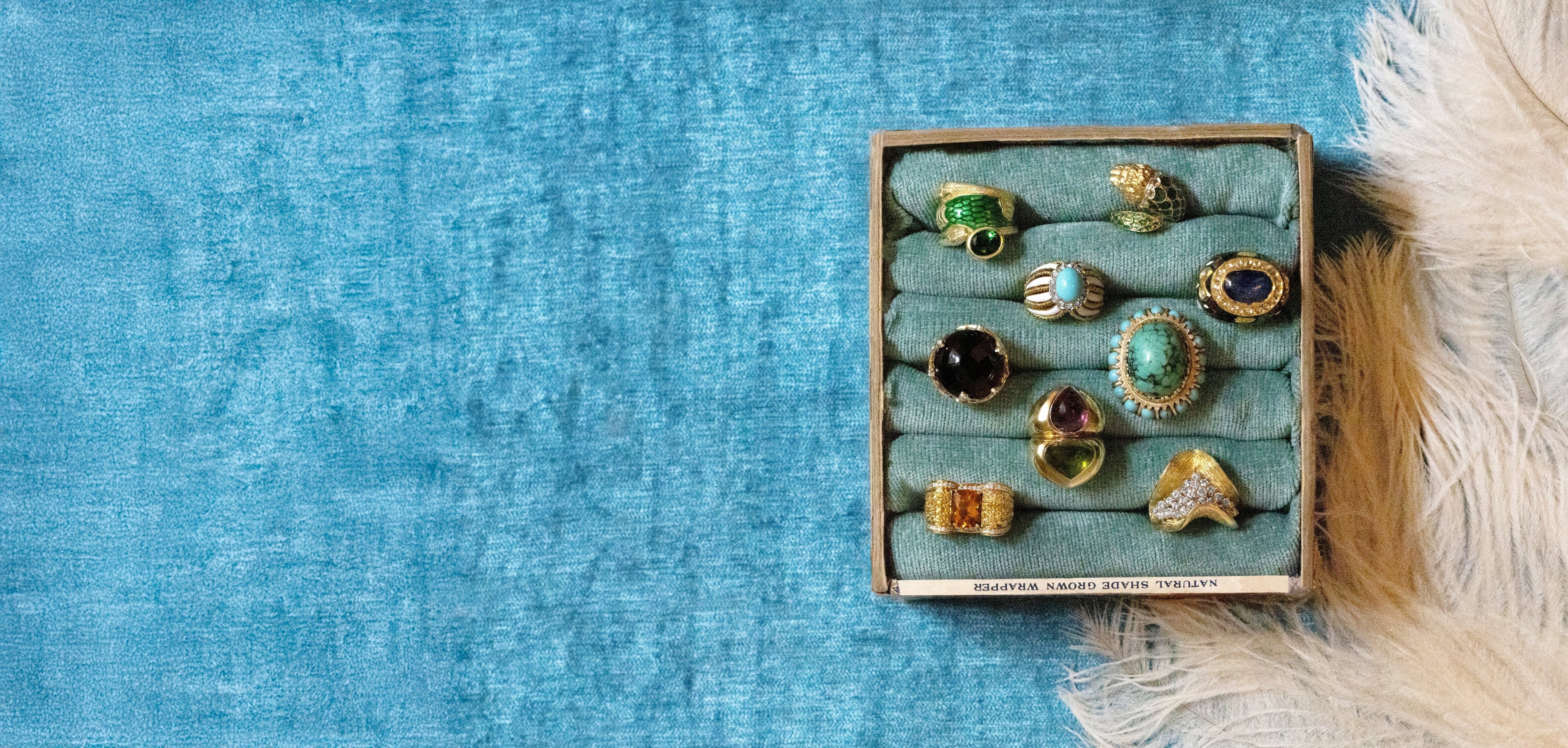 A vintage wooden ring box with a teal velvet interior, holding nine colorful rings, styled on a textured blue fabric background with a white feather on the right.
